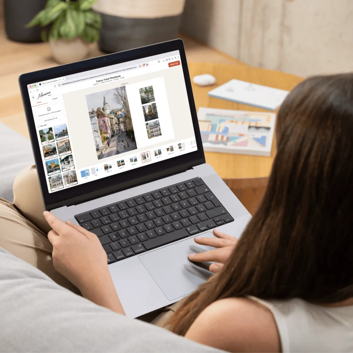 Someone laying out a France travel photobook in the Memorie Press editor on a laptop, with uploaded photos in the sidebar and a printed book on the table behind
